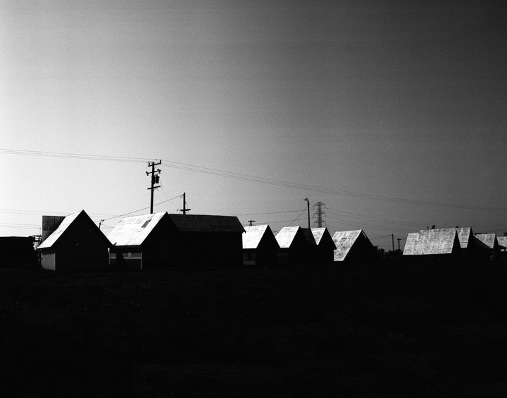 A row of small, dark buildings with bright roofs stands against a clear sky, with utility poles and power lines visible.