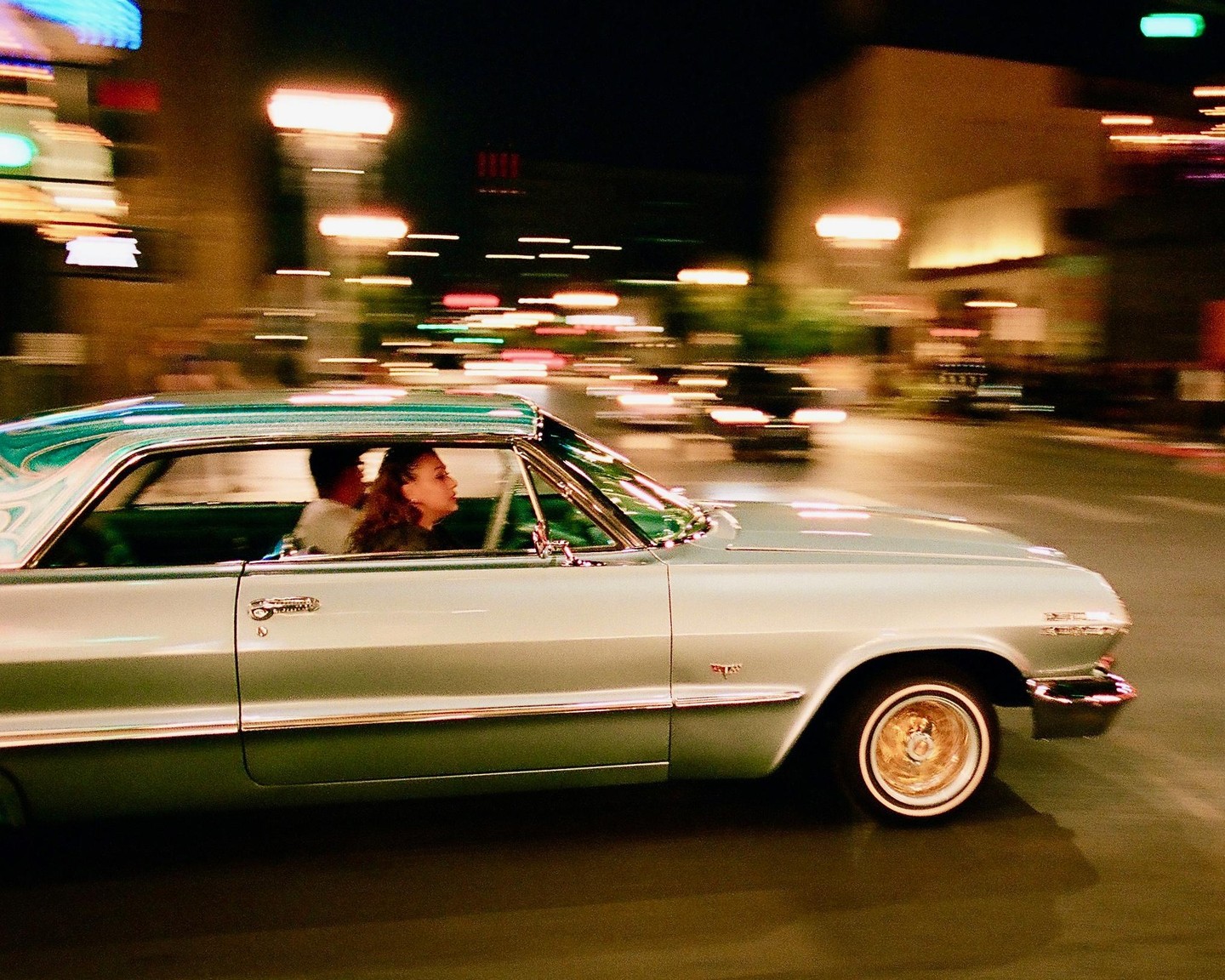 A woman rides in the passenger seat of a classic car at night, with blurred lights and buildings in the background.