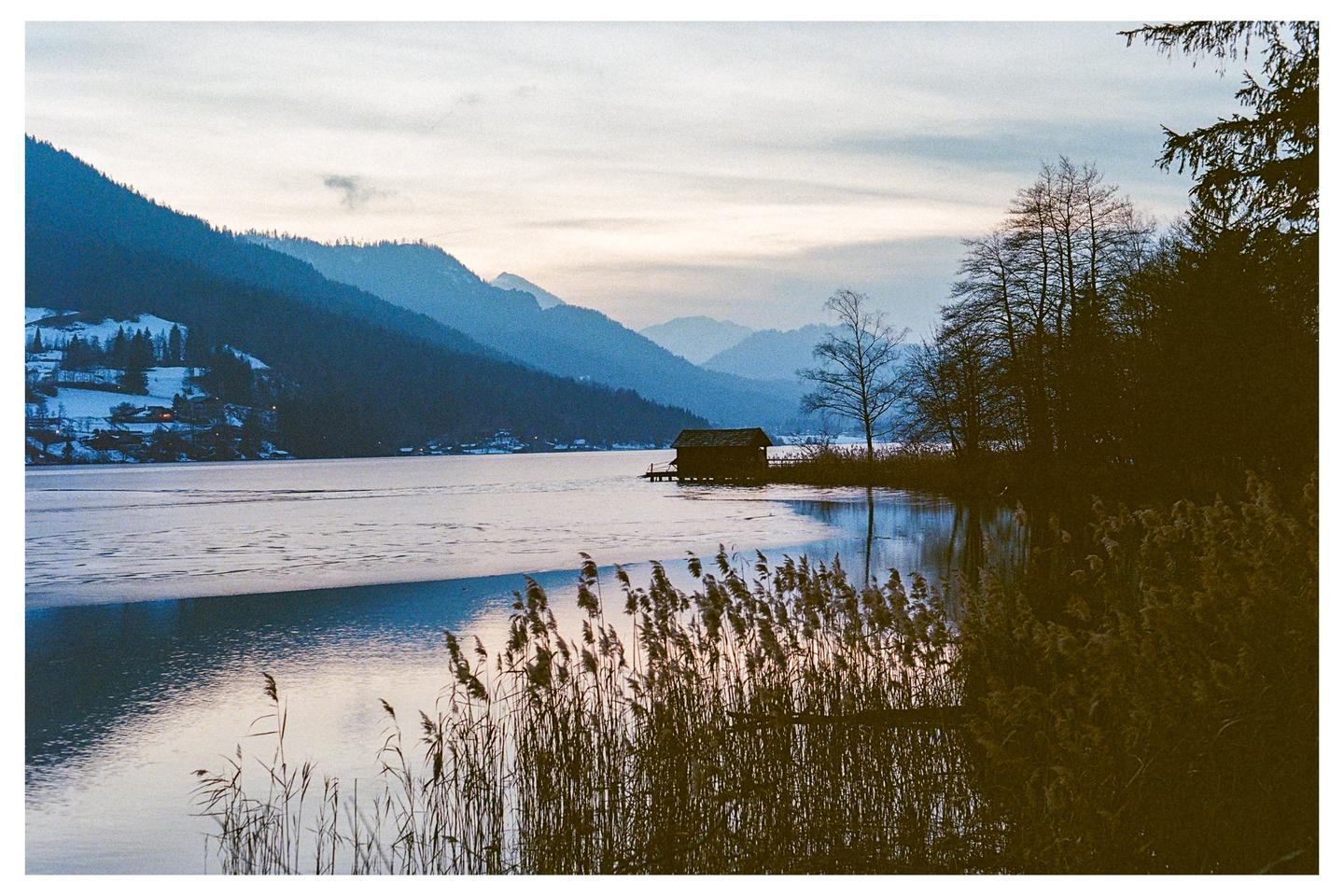 A small wooden cabin sits on the edge of a partially frozen lake, surrounded by mountains and reeds.