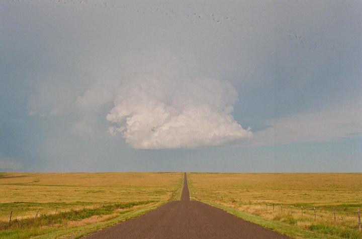 A long, straight road extends into the distance through a flat, grassy plain under a sky with a large cloud formation.