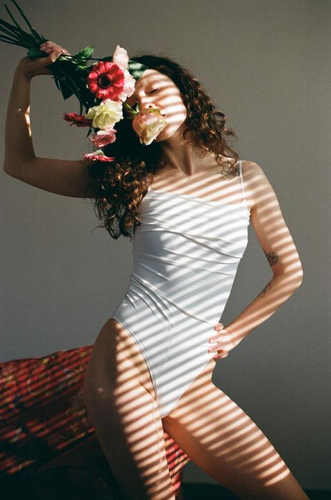 A woman in a white bodysuit holds a bouquet of flowers to her face, with shadows from blinds cast across her body.