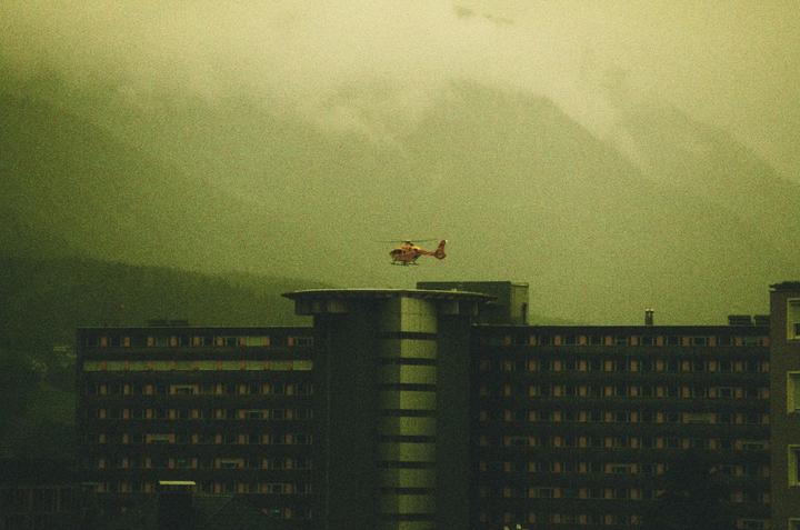 A red helicopter flies above a building with a helipad on its roof, with mountains in the hazy background.