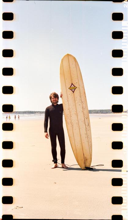 A man in a wetsuit stands on a sandy beach holding a surfboard.