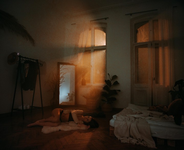 A woman lies on a rug on the floor of a dimly lit room with a bed and a clothing rack.
