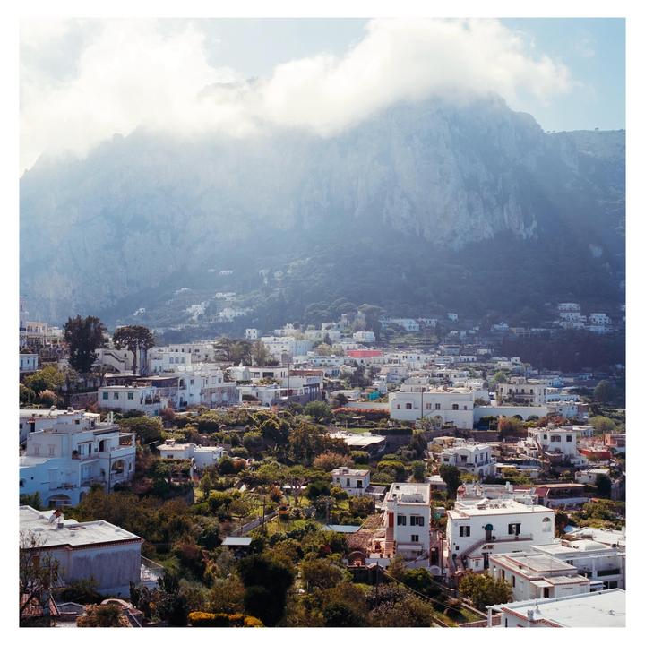A town with white buildings and lush greenery is nestled at the base of a large, cloud-covered mountain.