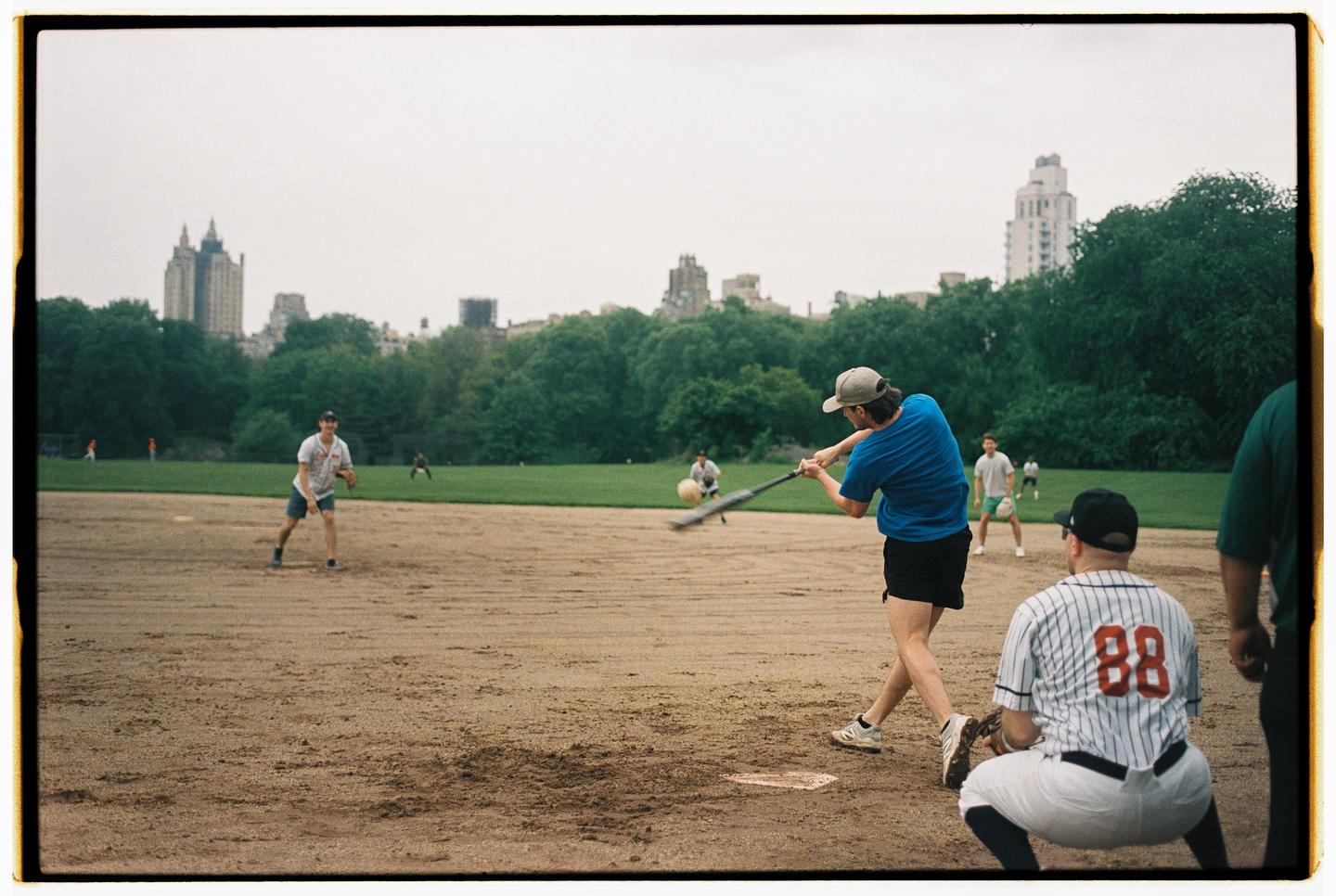 A man swings a baseball bat at a ball on a dirt field with buildings and trees in the background.