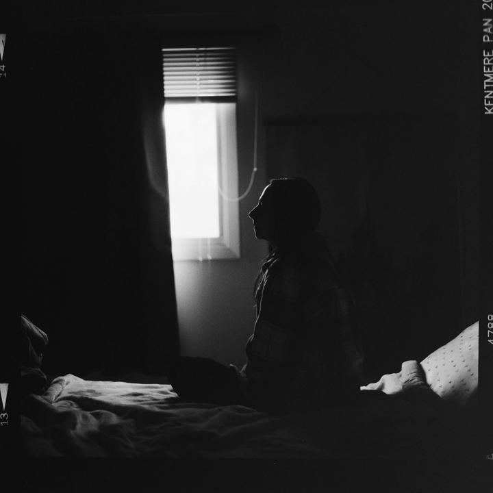 A woman sits on a bed in a dark room, looking towards a bright window with blinds.