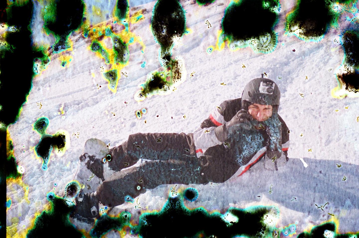 A man in winter clothing and a helmet is lying on a snowy slope, with colorful splotches and streaks across the image.