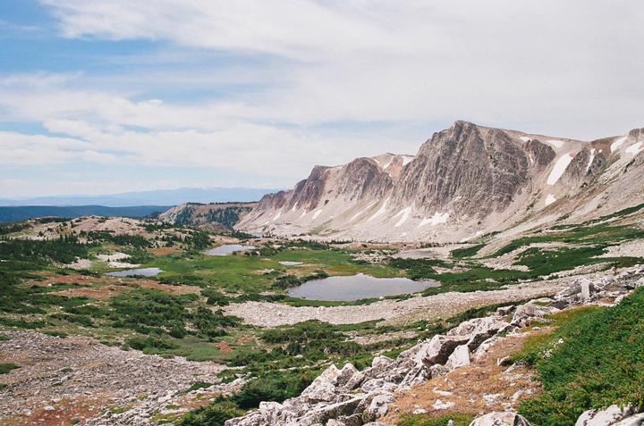 A vast mountain landscape features several small lakes nestled among rocky terrain and patches of green vegetation under a partly cloudy sky.