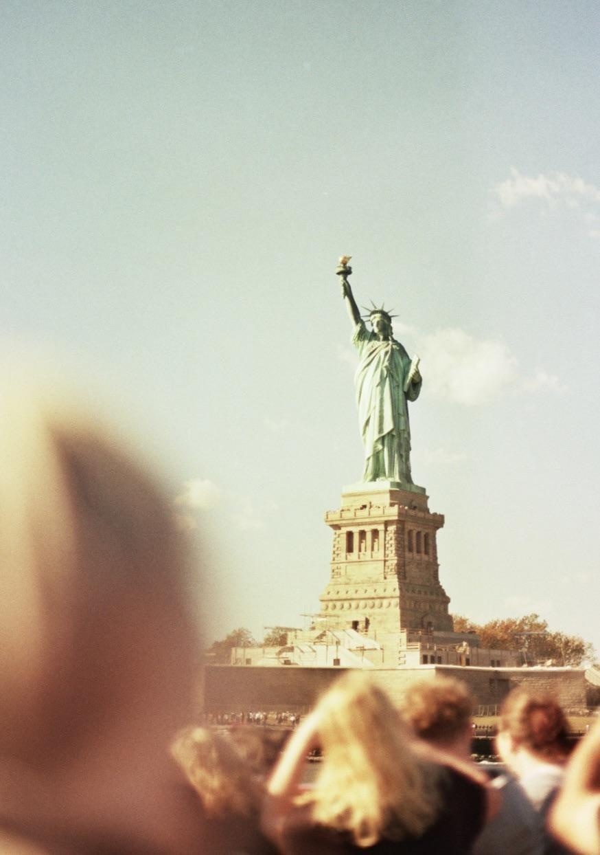 The Statue of Liberty stands tall against a clear sky, with the heads of people in the foreground.