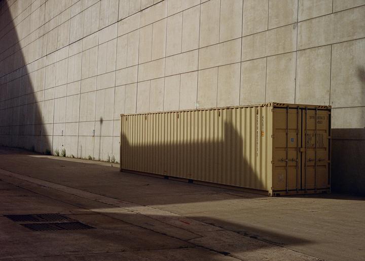 A long shipping container sits on a concrete surface next to a tall wall, with shadows cast across both.