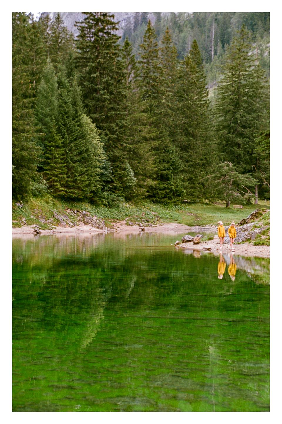 Two people in yellow jackets walk along the shore of a green lake surrounded by a forest.