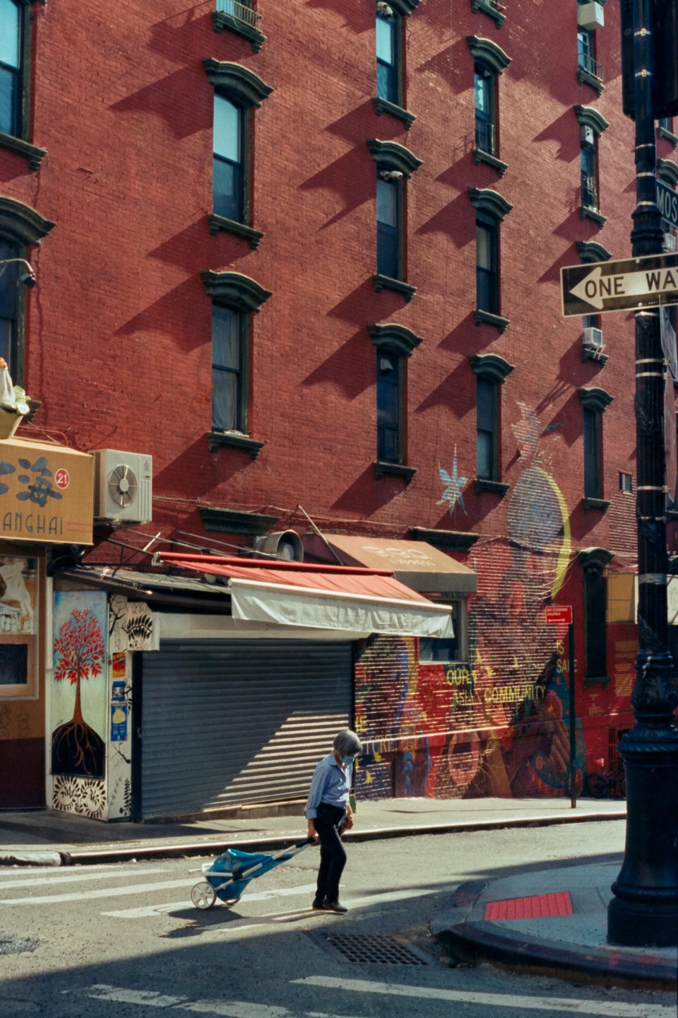 A person pulls a cart across a street in front of a red brick building with many windows and a mural.