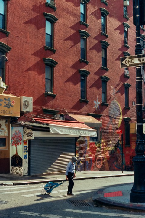 A person pulls a cart across a street in front of a red brick building with many windows and a mural.