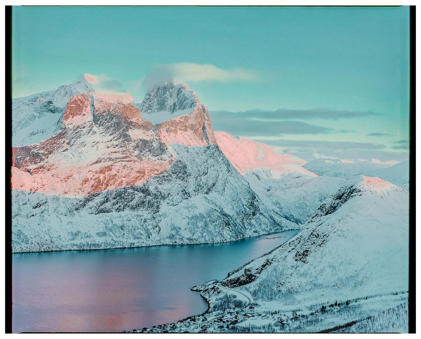 A snow-covered mountain range with a body of water and a small village at its base is illuminated by pink light.