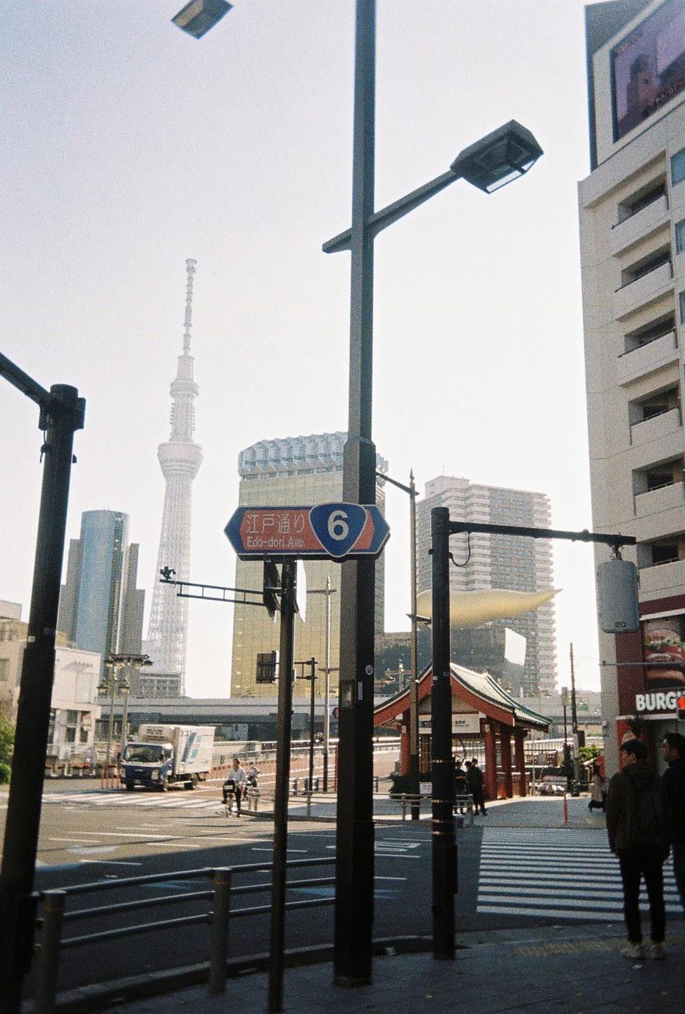 A street scene in Tokyo shows a tall tower in the distance, a street sign for Edo-dori Avenue, and people walking on the sidewalk.