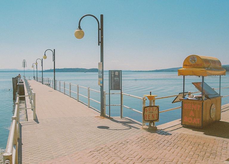 A long pier with streetlights extends into a calm lake under a clear sky, with a "rent a boat" stand on the right.