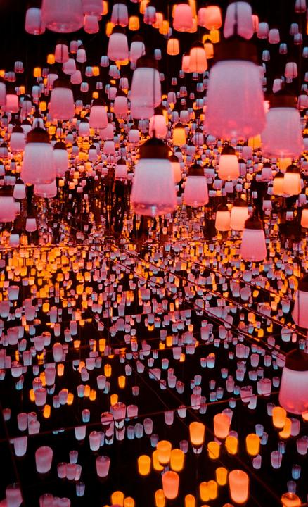Hundreds of glowing lanterns in shades of pink, orange, and white hang in a dark room, reflected in a mirrored surface.