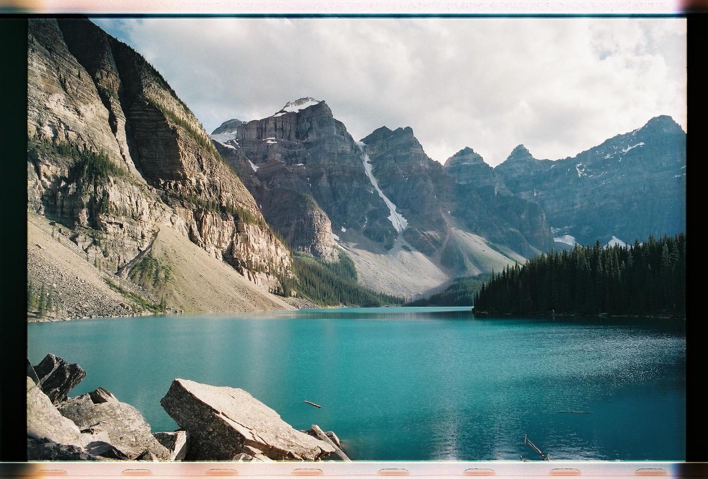 A turquoise lake is surrounded by mountains and a forest under a cloudy sky.