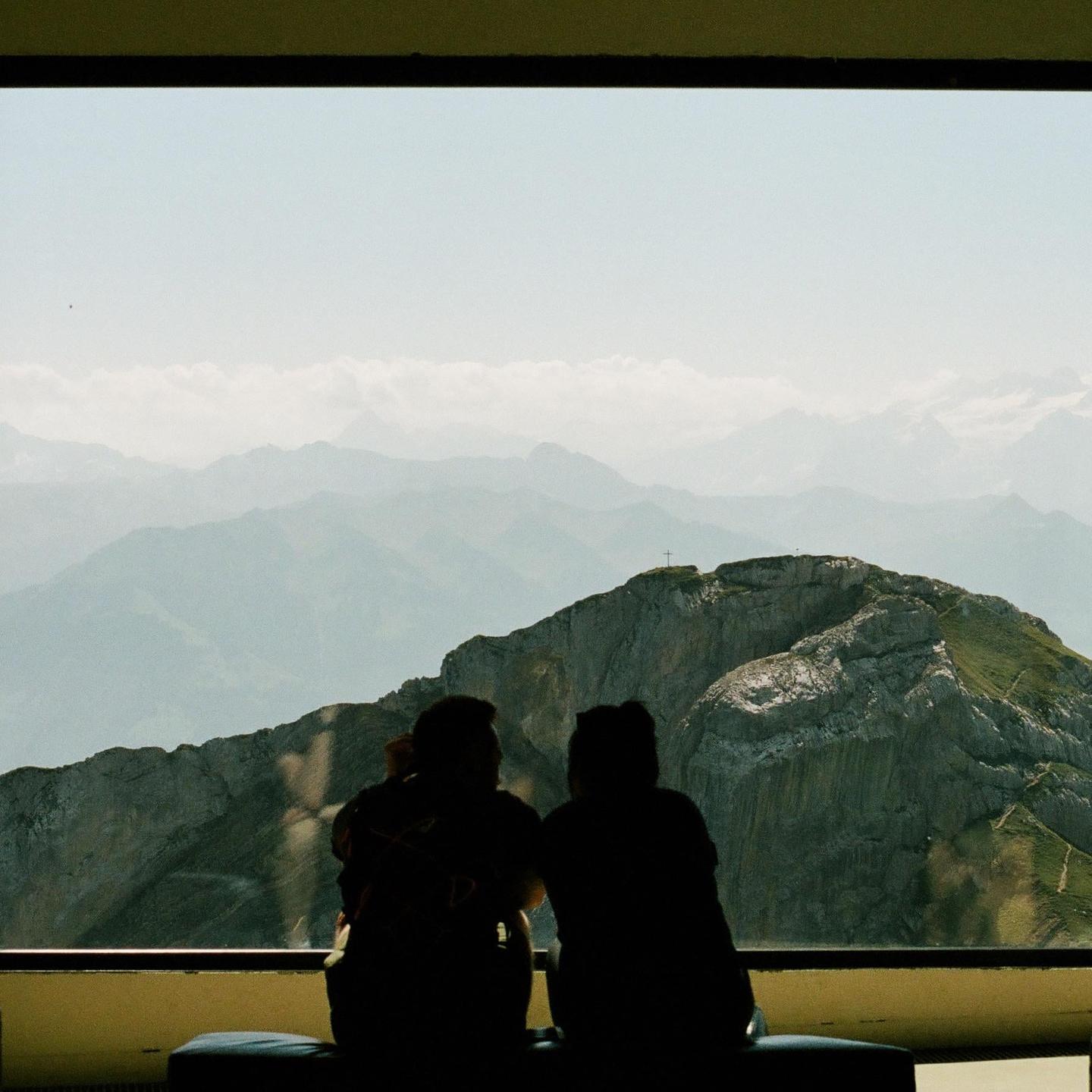 Two people sit with their backs to the viewer, looking out a window at a mountain range with a cross on the peak.