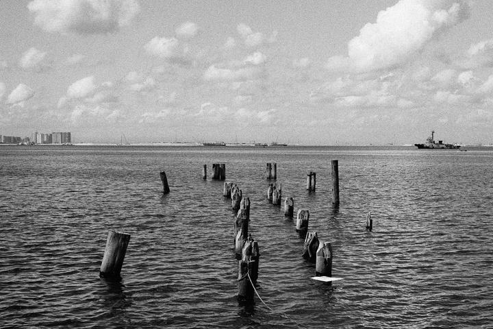 A body of water with wooden posts in the foreground and a city skyline and ship in the distance under a cloudy sky.