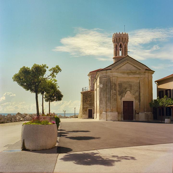 A church with a bell tower stands next to a paved area with trees and a planter, with the sea and a rocky shore in the background.