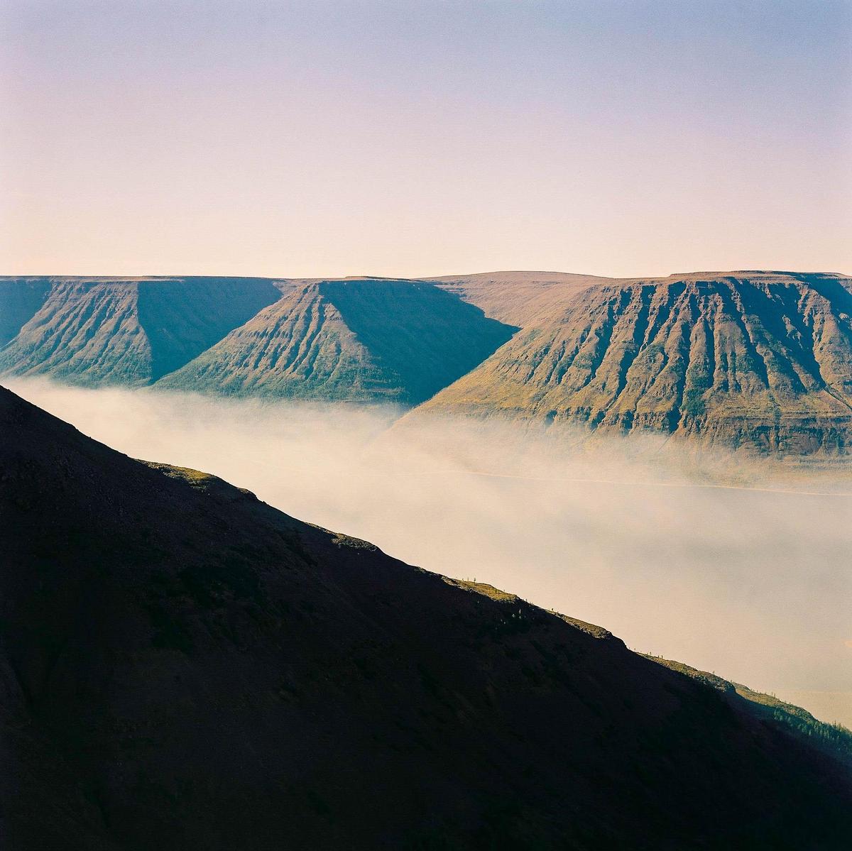 A layer of fog covers the valley between a dark mountain slope in the foreground and a range of mountains in the background.