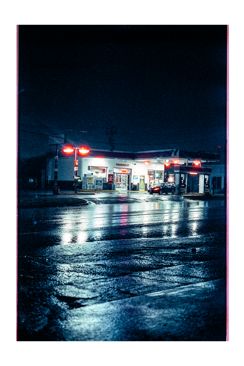A gas station and convenience store are illuminated at night, with reflections visible on the wet ground.