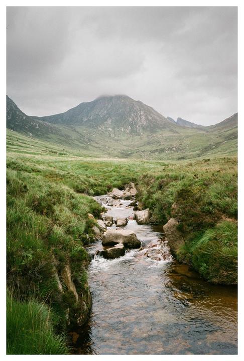 A stream flows over rocks through a grassy valley with mountains under a cloudy sky.