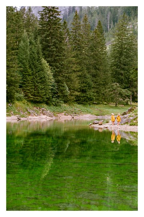 Two people in yellow jackets walk along the shore of a green lake surrounded by a forest.