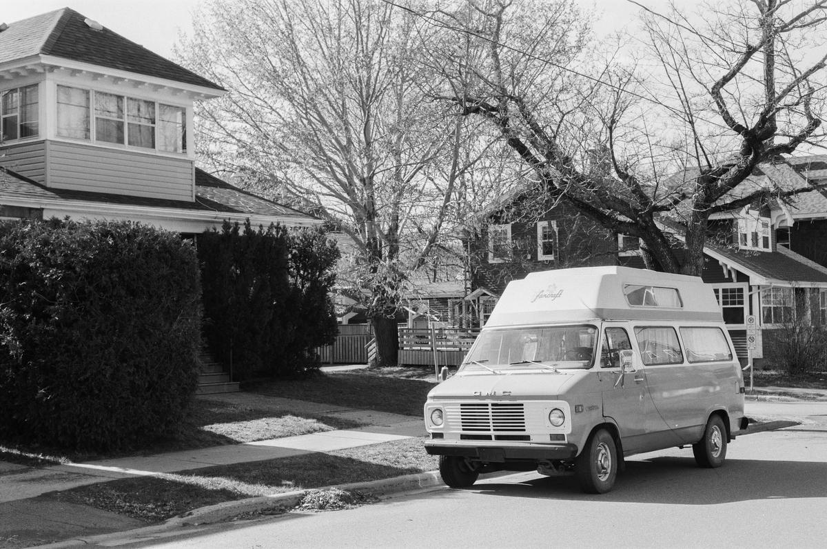 A camper van is parked on a residential street with houses and trees in the background.