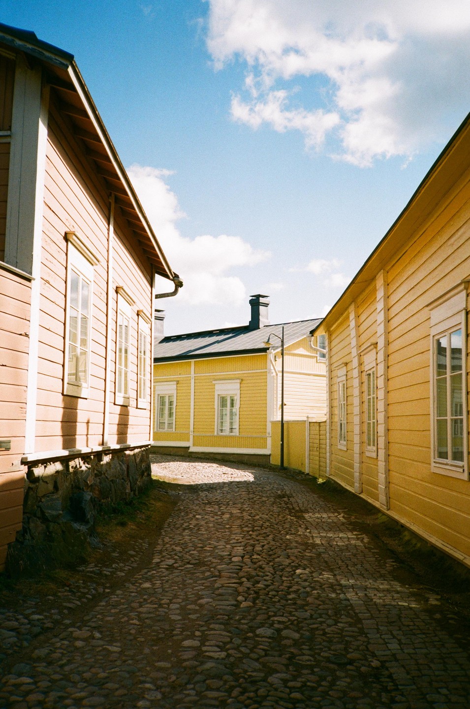A narrow cobblestone street is lined with yellow wooden buildings under a blue sky with white clouds.