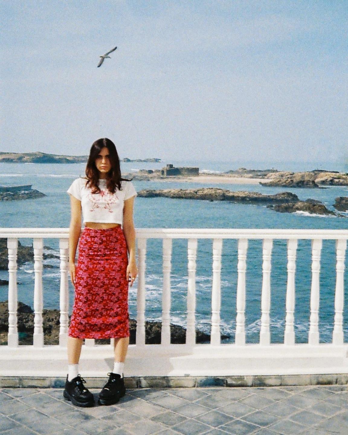 A woman stands on a balcony overlooking a rocky coastline with a bird flying overhead.