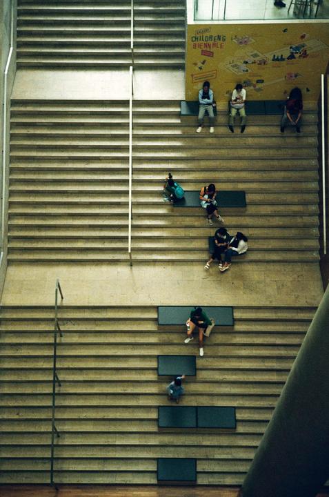 A high-angle view shows people sitting on a wide staircase with a yellow wall mural in the background.