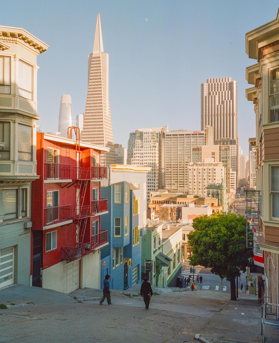 Two men walk down a steep street lined with colorful houses and tall buildings in the background.