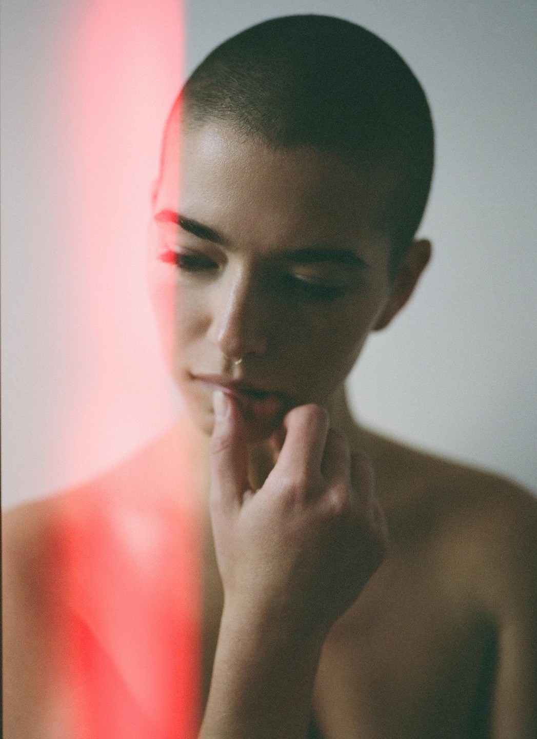 A person with a shaved head and a nose ring touches their lips with a finger, with a red light flare on the left side of the frame.