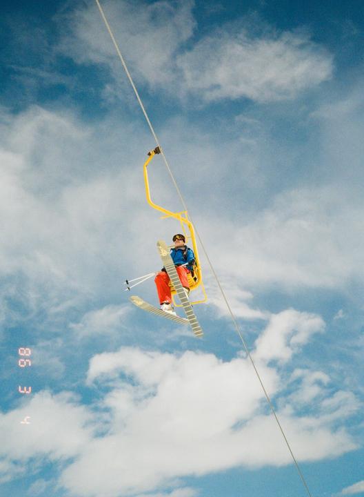 A person in ski gear is riding a yellow ski lift against a cloudy blue sky.