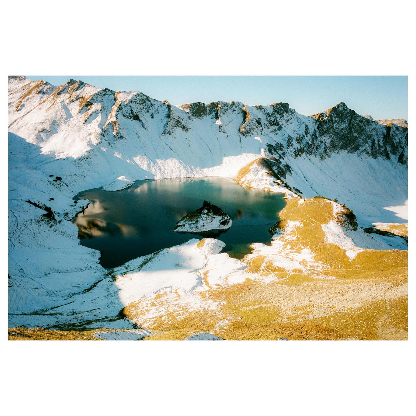 A high-angle landscape shows a lake surrounded by snow-covered mountains and patches of golden grass.