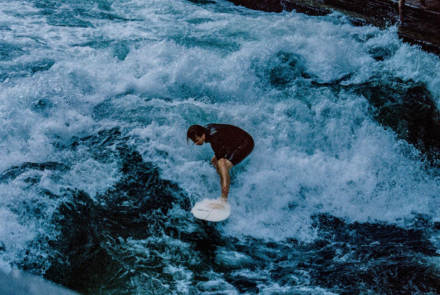 A man in a wetsuit surfs on a white board in turbulent water.