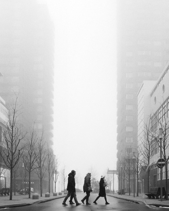 Three people walk across a street in front of tall buildings on a foggy day.