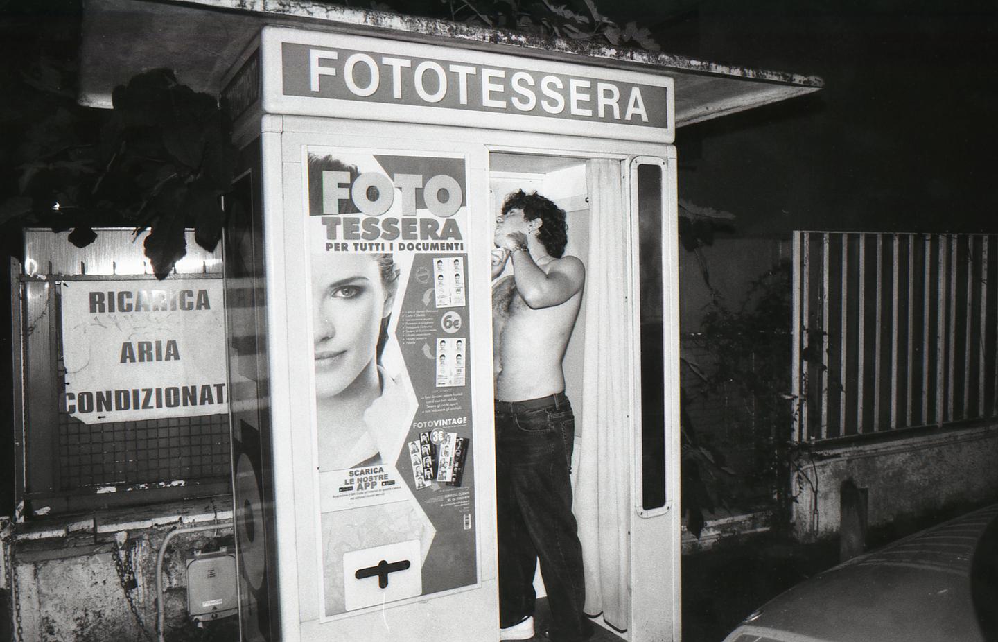 A shirtless man stands inside a photo booth at night, adjusting his hair.