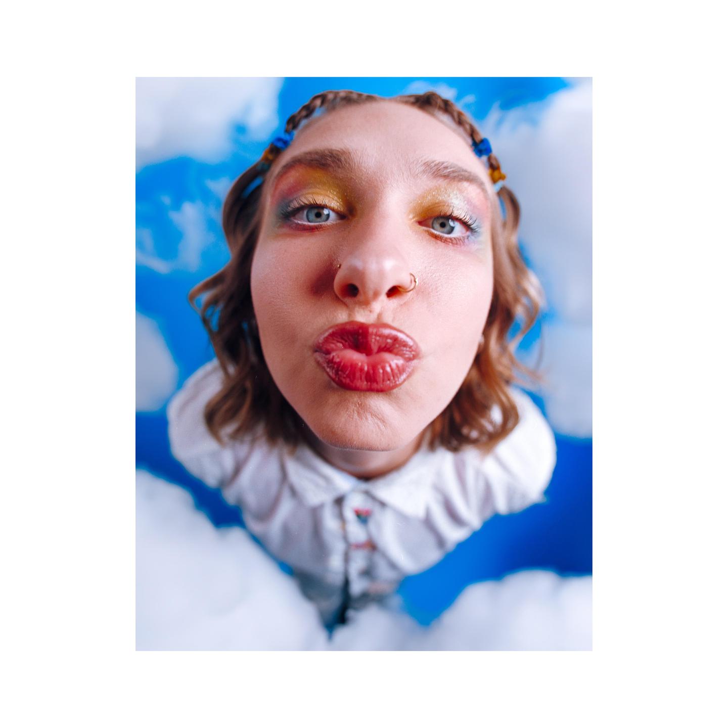 A woman with colorful eye makeup and braided hair puckers her lips for a kiss, surrounded by clouds against a blue sky.