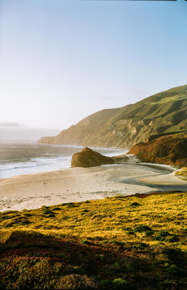 A sandy beach with a stream flowing into the ocean is nestled between a large rock formation and a grassy hillside under a bright sky.