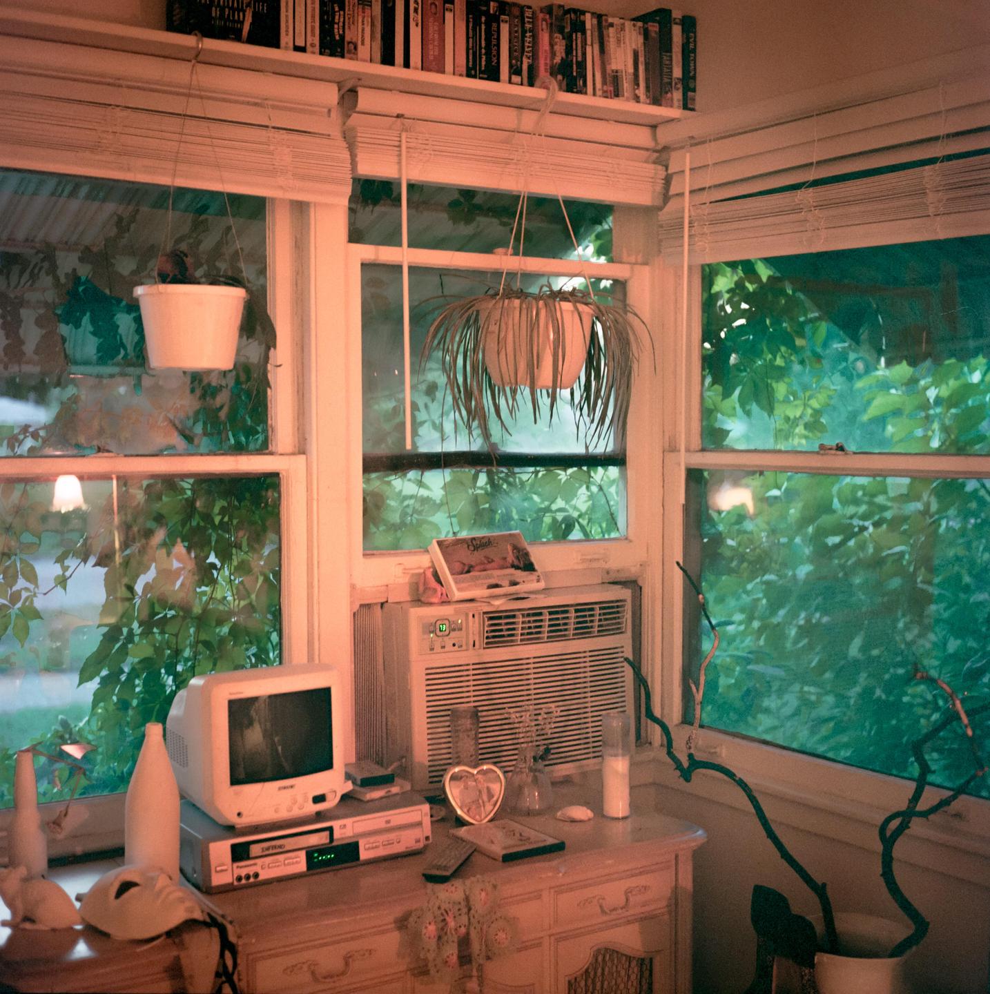 A corner of a room with windows, an air conditioner, a television, and various objects on a dresser.