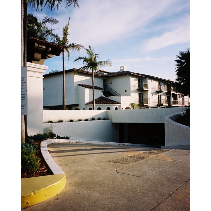 A white building with a red tile roof and palm trees is seen from a curved driveway leading to an underground garage.