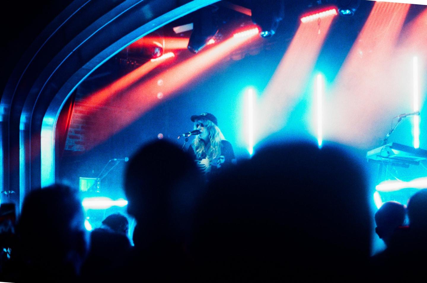 A woman with long hair and a cap sings into a microphone on a stage lit with red and blue lights, with the silhouettes of an audience in the foreground.