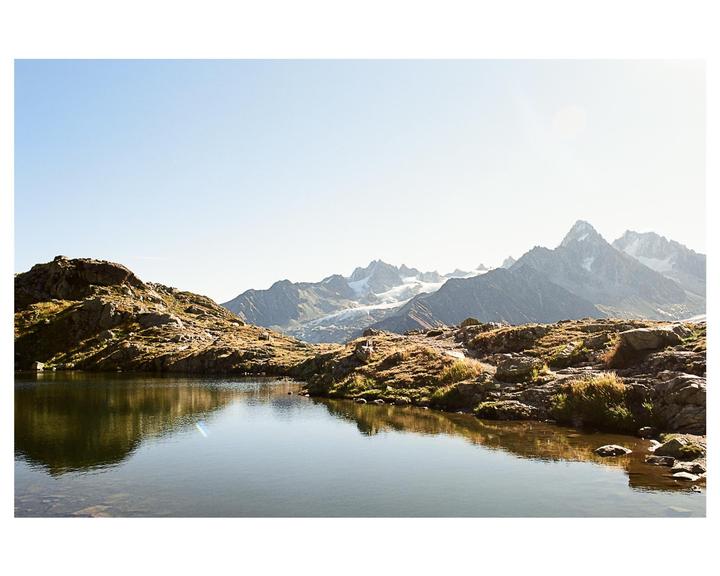 A clear mountain lake reflects the rocky shore and distant snow-capped peaks under a bright sky.