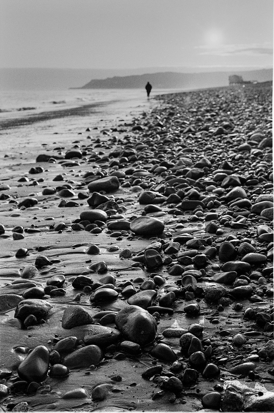 A person walks along a rocky beach with the sun low in the sky.