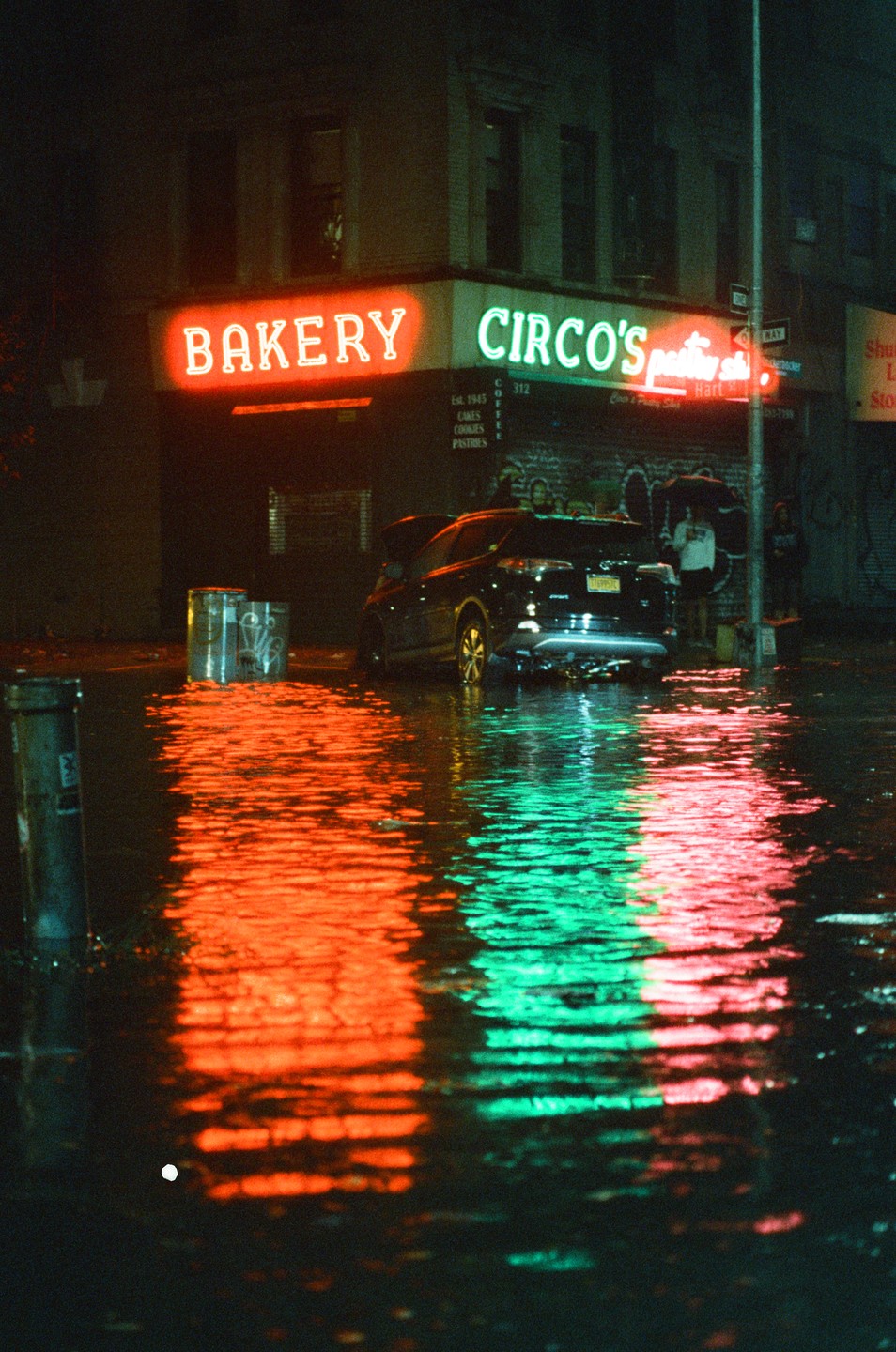 A street is flooded with water reflecting the neon signs of a bakery and a pastry shop at night.