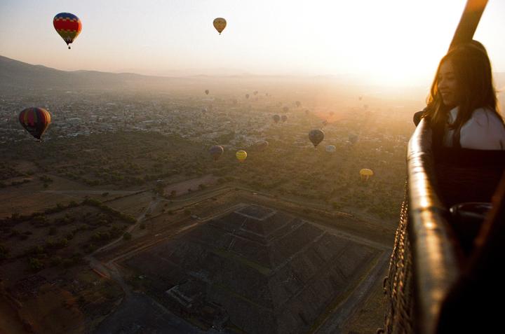 A woman looks out from a hot air balloon over a landscape with a pyramid and other hot air balloons at sunset.